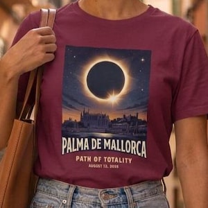 May include: A maroon short-sleeve t-shirt featuring a graphic of a solar eclipse over a city skyline. The text reads PALMA DE MALLORCA, PATH OF TOTALITY, AUGUST 12, 2026. The shirt is paired with light blue denim jeans and a brown leather shoulder bag.