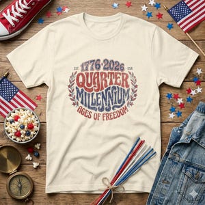 Peut inclure: Un t-shirt à manches courtes de couleur crème avec un motif vieilli rouge et bleu portant l'inscription 1776-2026 QUARTER MILLENNIUM AGES OF FREEDOM, entouré d'une couronne de laurier, centré sur le devant du vêtement.