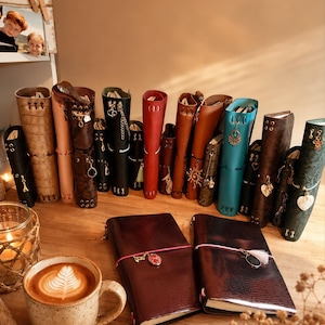 Puede incluir: Una colección de diarios encuadernados en cuero de varios colores, incluyendo negro, marrón, rojo, azul y verde, con dijes metálicos y cierres elásticos. Dos diarios de color marrón oscuro yacen planos junto a una taza de café.