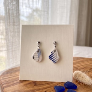 Op de afbeelding: Een paar keramische oorhangers. Elke oorbel heeft een unieke vorm met een witte basis en blauwe gestreepte en patroonontwerpen. De oorbellen worden gepresenteerd op een crèmekleurige kaart, met blauwe glazen kiezels en een gedroogde plant.