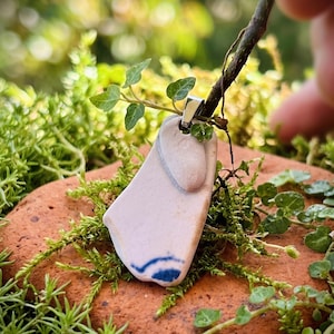 May include: A handmade pendant necklace featuring a piece of light pink and white ceramic with a blue design. The pendant is suspended from a silver-colored bail and is displayed on a bed of green moss and a red-brown stone.