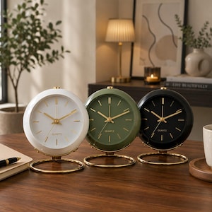 May include: Three round quartz clocks in white, olive green, and black, each with gold-colored hands and a gold base. The clocks are displayed on a wooden surface, with a pen and notebook visible in the foreground. The clock faces have the word "QUARTZ" printed on them.