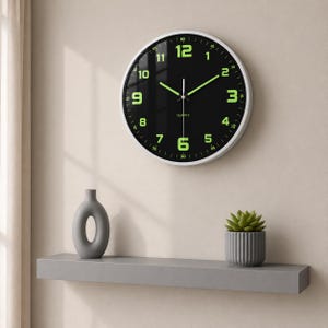 Puede incluir: Un reloj de pared redondo con marco blanco, esfera negra y números verdes. El reloj está montado en una pared beige. Debajo, una estantería gris con un jarrón gris y una pequeña suculenta en maceta.