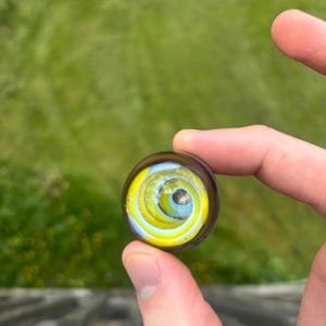 May include: A glass marble with a spiral design in yellow, light blue, and brown. The marble is held between fingers against a blurred green background.