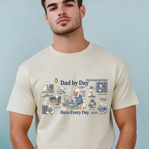 May include: Beige t-shirt with a graphic featuring cartoon ducks in various tech-related scenarios. Text includes "Dad by Day," "Tech Dad Edition," and "Hero Every Day." The design incorporates computer screens and other tech elements.