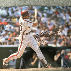 Può includere: Fotografia firmata di un giocatore di baseball in uniforme dei Phillies, a metà swing con una mazza. Il giocatore indossa un casco rosso, una divisa a righe bianche e rosse e tacchetti marroni. Lo sfondo mostra una folla sfocata.