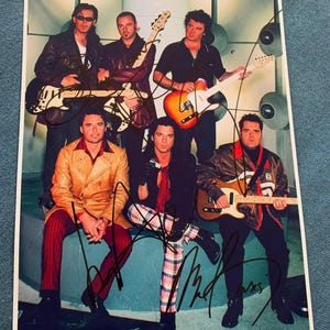 May include: A photograph of a band posing with guitars. The band members are wearing various outfits, including leather jackets, button-down shirts, and patterned pants. The image is signed by the band members.