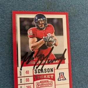 Può includere: Una carta collezionabile di football rossa e bianca con un giocatore in uniforme e casco rossi. La carta include il testo "SEASON TICKET" e "CONTENDERS 20 DRAFT PICKS 17". La carta è firmata con inchiostro nero.
