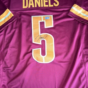 May include: Maroon football jersey with the name "DANIELS" in yellow block letters across the shoulders. The number "5" is prominently displayed on the front, outlined in white with a yellow and orange dotted pattern. The jersey has yellow and orange stripes on the sleeves.
