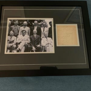 May include: Framed black and white photograph of a group of men in suits, with a smaller framed document to the right. The photograph is matted and framed in a black frame.
