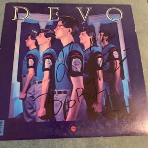 Op de afbeelding: Albumhoes voor Devo's album "New Traditionalists". De hoes toont vijf bandleden in profiel, gekleed in blauwe shirts en grijze broeken. De albumtitel staat bovenaan. Handtekeningen van de bandleden zijn zichtbaar.