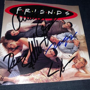 Puede incluir: Un póster de Friends con el logo del programa y una foto grupal del elenco acostado en una cama. El póster tiene múltiples firmas en tinta negra en la imagen.