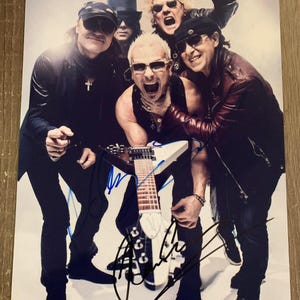 May include: A photograph of a rock band posing with a guitar. The band members are wearing sunglasses, leather jackets, and hats. The image is signed by the band members. The photo has a white background and a "Real Authentic" stamp.