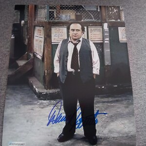 May include: A rectangular photograph featuring a person wearing a light-colored button-down shirt, a dark vest, a dark tie, and dark trousers. The image includes a blue ink signature across the lower portion and an Authentication Direct sticker.