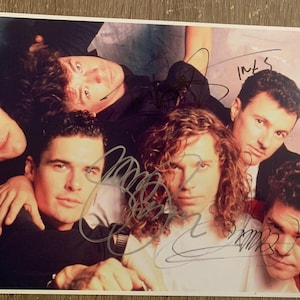 May include: A color photograph of a group of six men, likely a band, posing closely together. The image is signed by the individuals. The men are wearing dark clothing and sunglasses. The image has a "Real Authentic" sticker.