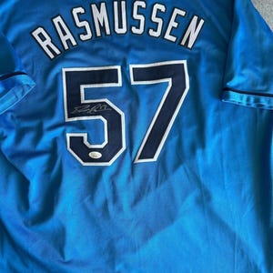 May include: Light blue baseball jersey with the name "RASMUSSEN" in white lettering above the number "57" in navy blue with a white outline. The jersey has a signature below the number.