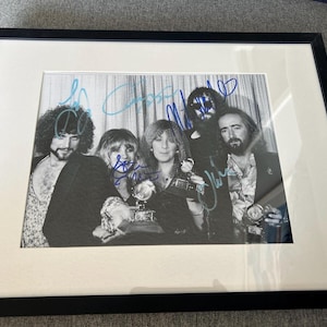 May include: Framed black and white photograph of a band posing with awards. The image is signed in blue ink. The photograph is set in a white mat and a black frame.