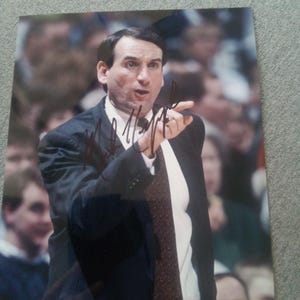 May include: A glossy photograph of a basketball coach wearing a dark suit, white shirt, and patterned tie, pointing forward with an open mouth. The image features a dark signature across the center and a small authentication sticker in the corner.