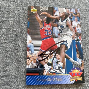 May include: A basketball trading card depicting a game scene with two players. One player is in a red jersey with the number 33, the other in a blue and white jersey with the number 32. The card is signed and has the text "NBA Top Prospects".