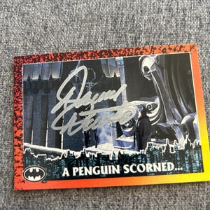 May include: A Batman trading card with a scene featuring the Penguin. The card has a red and orange border and the text "A PENGUIN SCORNED..." along the bottom. The card is signed in silver ink.