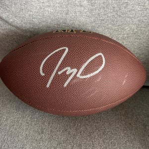 May include: A brown leather American football with white stitching and a white signature. The ball has a textured surface. The NFL logo is visible. Designed for American football games.