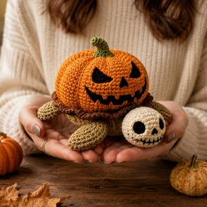May include: A crocheted turtle toy featuring a bright orange pumpkin shell with a dark brown ruffled edge and a carved jack-o'-lantern face. The turtle has a light beige head with a stitched skull design, dark eye sockets, and a stitched mouth.
