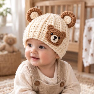 Puede incluir: Un gorro de ganchillo color crema con dos orejas de oso redondas en marrón y crema en la parte superior y una aplicación de cara de oso marrón en el lateral. El gorro tiene una textura acanalada e incluye pequeños ojos negros y una nariz bordada.