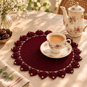May include: A round, deep burgundy crochet placemat featuring a scalloped border of heart-shaped motifs accented with small gold beads. The placemat is crafted with a dense, circular stitch pattern and serves as a base for a floral teacup and saucer.