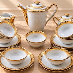 Op de afbeelding: Een theeservies van wit porselein bestaande uit een hoge theepot, een suikerpot met deksel, een melkkan en meerdere theekopjes met bijpassende schoteltjes. Elk onderdeel is versierd met een brede, gouden filigraanrand langs de randen.