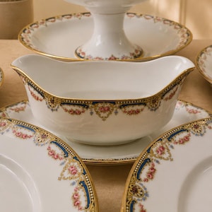 Peut inclure: Une saucière et une coupe sur pied en porcelaine blanche ornées d'une bordure décorative de roses roses, de volutes bleues et d'un liseré doré. L'ensemble comprend des assiettes blanches assorties avec le même motif floral et doré.