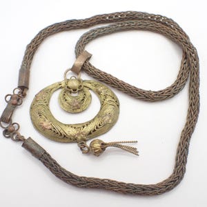 May include: An antique brass-toned necklace with a woven chain and a large, openwork pendant. The pendant features a circular design with floral accents and a small tassel. The necklace is displayed on a white background.