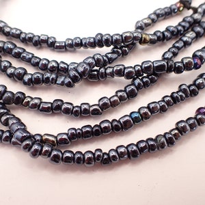 Peut inclure: Gros plan de plusieurs brins de perles gris foncé, iridescentes. Les perles sont petites, de forme carrée et ont un éclat métallique. Les brins sont disposés en couches, créant un effet texturé. Les perles reflètent des reflets violets, bleus et dorés.