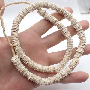 May include: A necklace made of small, flat, beige-colored beads strung together on a natural-colored cord. The beads are uniform in size and shape, creating a circular pattern. The necklace is held in a hand against a white background.