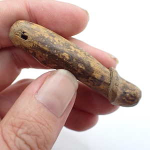 Könnte beinhalten: Ein kleines, handgeschnitztes Holzobjekt mit zylindrischem Körper und abgerundetem Ende. Das Holz hat ein natürliches, gealtertes Aussehen mit dunkelbraunen und beigen Farbtönen. Ein kleines Loch ist oben gebohrt.