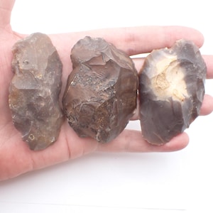 May include: Three knapped flint stones with irregular, sharp-edged surfaces and earthy brown and grey tones. Each stone displays distinct conchoidal fractures and a textured, chipped appearance characteristic of hand-worked lithic tools.