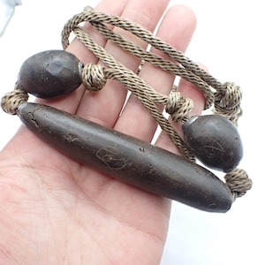 Puede incluir: Un collar marrón oscuro con dos cuentas redondeadas y una cuenta larga y cilíndrica. Las cuentas están conectadas por un cordón tejido de color beige. El collar se sostiene en una mano.