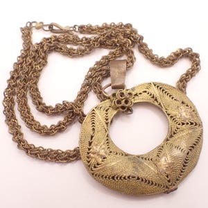 Può includere: Una collana con pendente vintage in filigrana dorata. Il pendente è un design traforato circolare con accenti floreali. La catena è in tinta oro abbinata e ha una chiusura a moschettone. La collana è un gioiello importante.