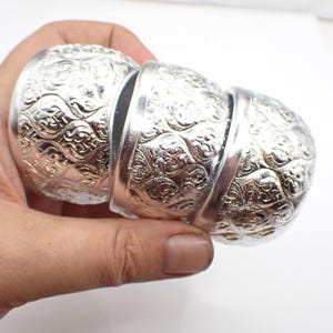 May include: Three silver bangles with intricate floral designs. The bangles are stacked on top of each other and are made of a shiny metal.