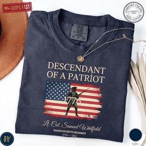 Pode incluir: Uma t-shirt azul-marinho de manga curta com o texto "DESCENDANT OF A PATRIOT" sobre uma bandeira americana desgastada, um soldado com uma espingarda e o ano "1776". Abaixo, "Lt. Col. Samuel Whitfield" e "MASSACHUSETTS REGIMENT 1776-1781".