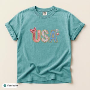 Può includere: Una t-shirt a maniche corte color verde schiuma di mare con la scritta USA in un carattere decorativo. La lettera U ha un motivo a quadretti rossi e bianchi con un fiocco, la S una stampa floreale e la A un motivo a stelle blu e bianche.