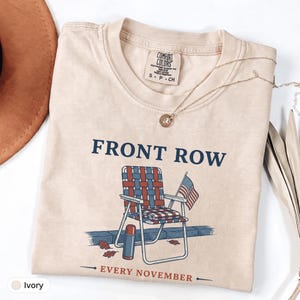 Peut inclure: Un t-shirt à manches courtes ivoire avec un motif représentant une chaise de jardin tressée, un petit drapeau américain et un thermos. Le texte FRONT ROW est inscrit au-dessus et EVERY NOVEMBER en dessous. Étiquette Comfort Colors.