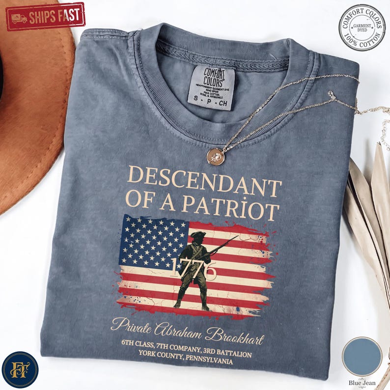 Pode incluir: Uma t-shirt azul ganga com o texto DESCENDANT OF A PATRIOT sobre um gr&aacute;fico da bandeira americana com a silhueta de um soldado e o ano 1776. Abaixo da bandeira est&aacute; o texto Private Abraham Brookhart e detalhes da unidade militar.