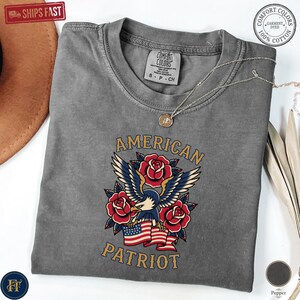 May include: A charcoal grey short-sleeve t-shirt featuring a graphic of a bald eagle with spread wings, red roses, and an American flag. The text reads AMERICAN PATRIOT. The shirt has a crew neckline and a Comfort Colors brand label inside.