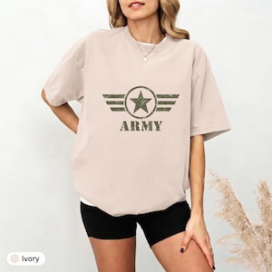 Pode incluir: Uma t-shirt de manga curta na cor marfim com um gráfico verde-oliva desgastado no peito. O design apresenta uma estrela central dentro de um círculo com asas horizontais em cada lado, acima da palavra ARMY em uma fonte de bloco em negrito.