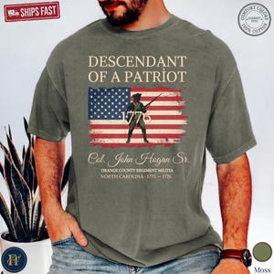 Pode incluir: Uma t-shirt de manga curta verde musgo com um gr&aacute;fico da bandeira americana e um soldado segurando um rifle. O texto diz: DESCENDANT OF A PATRIOT, 1776, Col. John Hogan Sr., ORANGE COUNTY REGIMENT MILITIA, NORTH CAROLINA, 1775-1776.