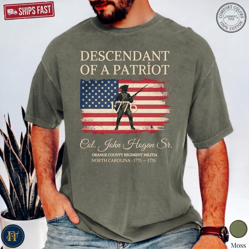 Pode incluir: Uma t-shirt de manga curta verde musgo com um gr&aacute;fico da bandeira americana e um soldado segurando um rifle. O texto diz: DESCENDANT OF A PATRIOT, 1776, Col. John Hogan Sr., ORANGE COUNTY REGIMENT MILITIA, NORTH CAROLINA, 1775-1776.
