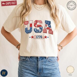 May include: An ivory short-sleeve t-shirt featuring the letters USA in a patchwork design of red, white, and blue patterns including gingham, stars, and florals. Below the letters is a rectangular badge with the text 1776 flanked by two red stars.