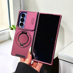 Puede incluir: Una funda para smartphone plegable de color rosa oscuro con una cubierta deslizante para la lente de la cámara, un soporte de anillo metálico circular en la parte posterior y un acabado mate. Diseñada para un dispositivo con pantalla plegable.