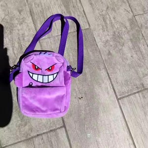 May include: A plush purple crossbody bag featuring a character face with red eyes and a wide white grin. The bag has a front pocket, a top zipper closure, and a matching purple adjustable shoulder strap attached with silver-toned metal hardware.