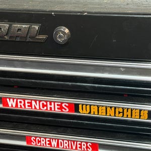 May include: A black metal tool chest drawer unit featuring silver trim and a circular key lock. The drawers are labeled with red and white rectangular stickers reading WRENCHES, SCREWDRIVERS, and SPECIALTY, alongside a yellow WRENCHES decal.